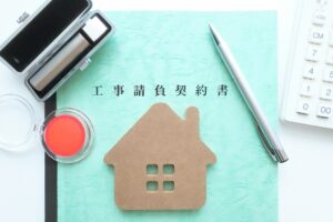 Teal document titled 工事請負契約書 with a pen, stamp pad, and a cardboard house cutout beside a calculator.