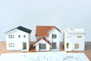 Row of white architectural model houses with brown and black roofs on a wooden table, blueprints underneath.