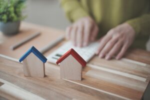 Two wooden toy houses with blue and red roofs sit on a wooden table, blurred hands typing on a calculator in the background.