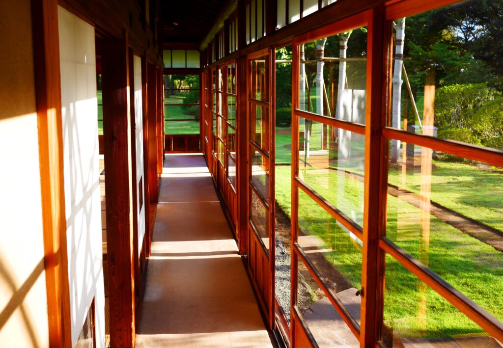 Sunlit wooden hallway with large glass windows overlooking a green lawn and trees outside.