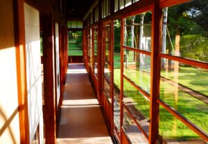 Sunlit wooden hallway with large glass windows overlooking a green lawn and trees outside.