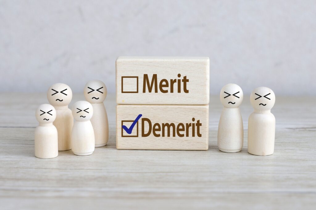 Block stack comparing Merit and Demerit with a checked Demerit box, surrounded by small wooden figures with distressed faces, conveying evaluation or judgment.