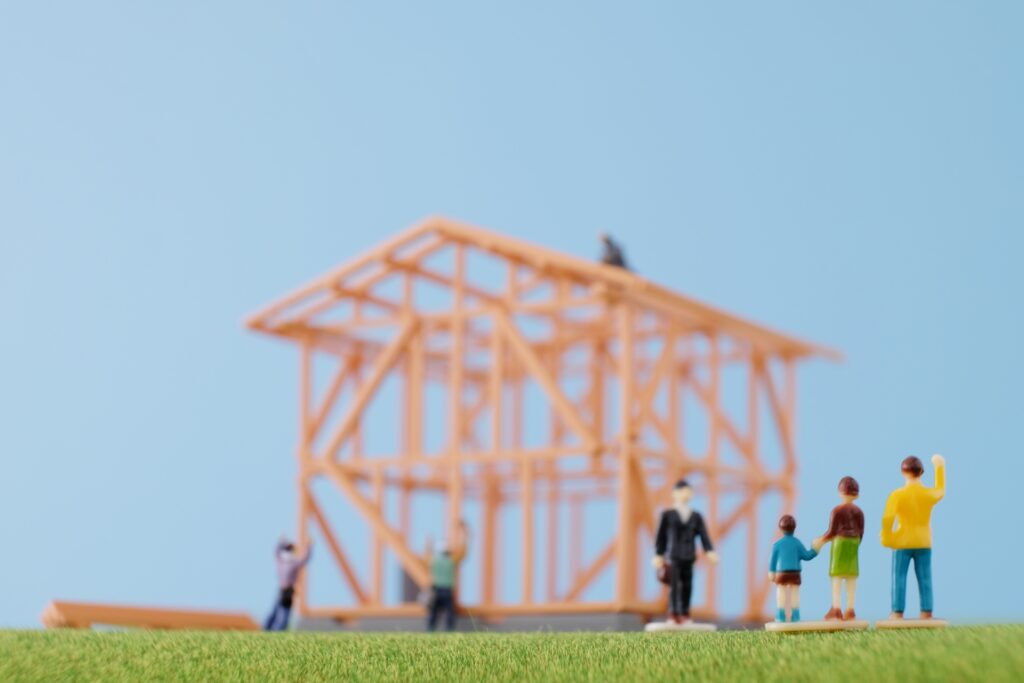Toy construction site scene with an orange steel framework in the background and miniature workers standing on a grassy surface under a blue sky.