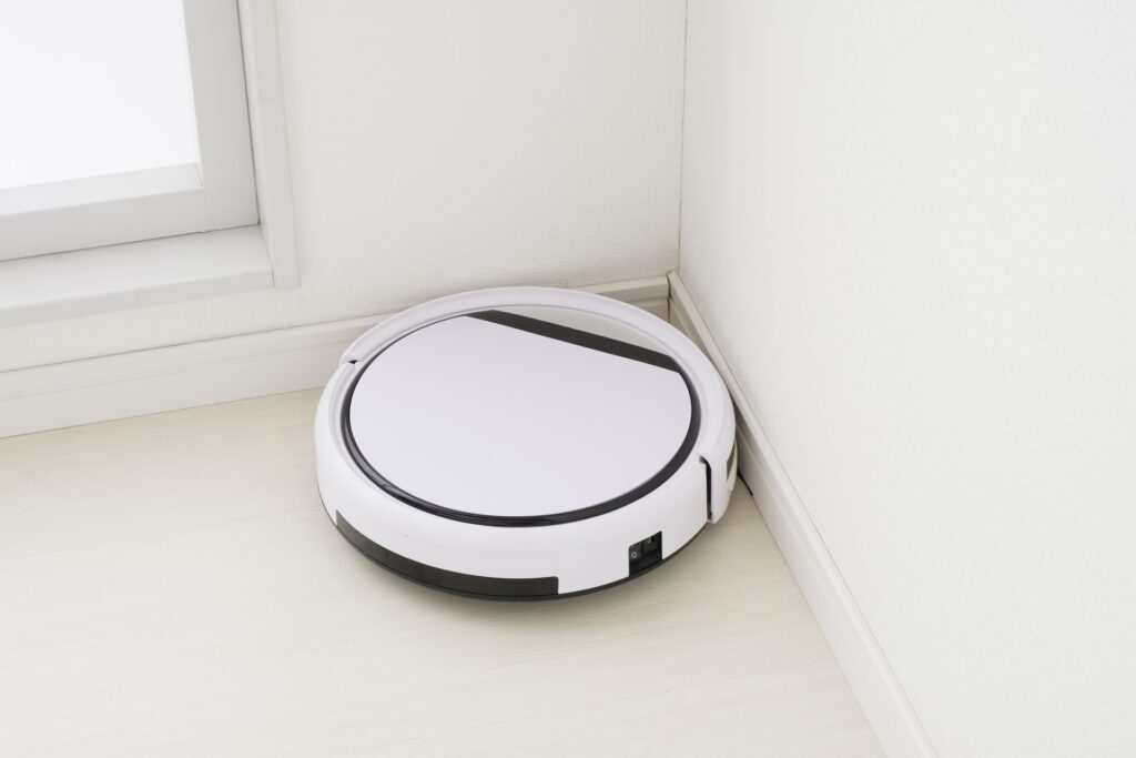 Round white robotic vacuum resting in a corner by the baseboard and window on a light floor.