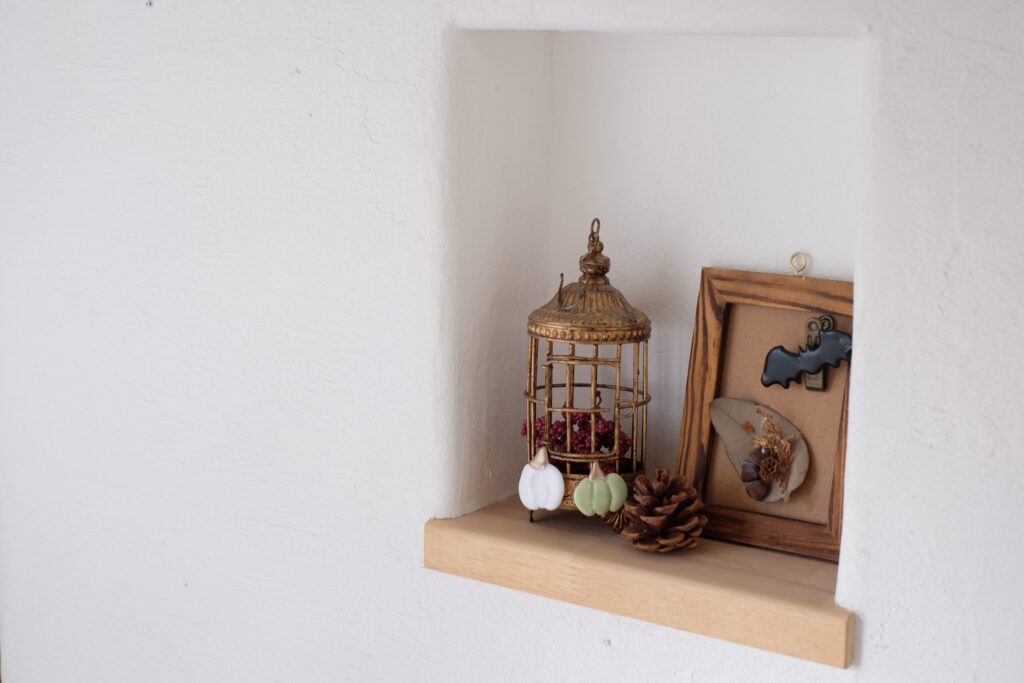 Decorative gold birdcage and framed botanical display on a wooden shelf against a white wall, with pine cones and small bulb ornaments nearby.