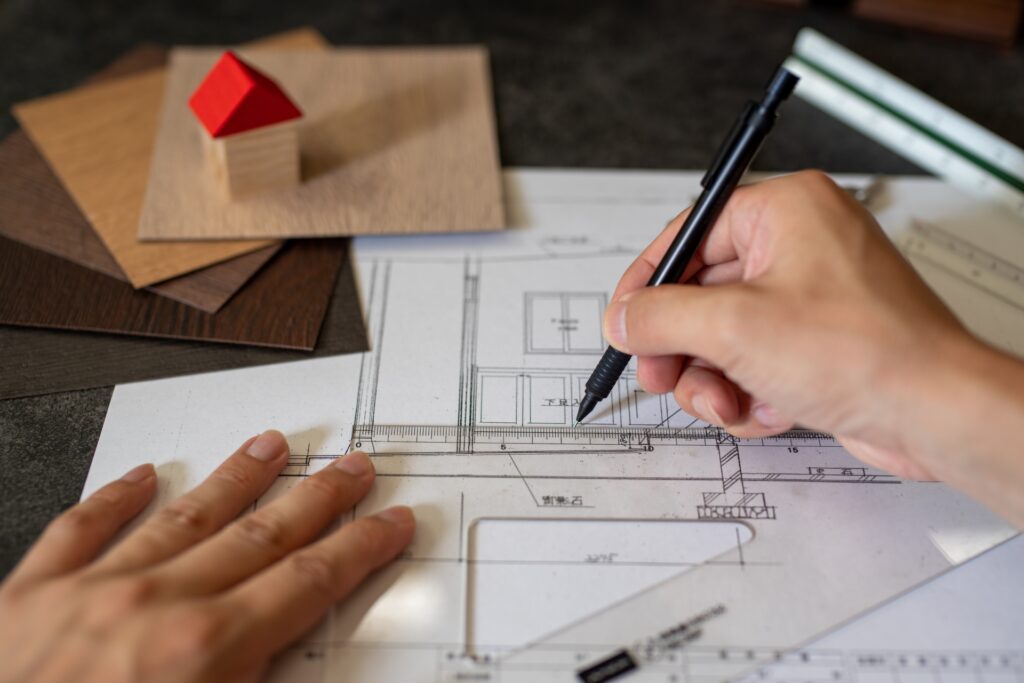 Hands drafting architectural plans on blueprints with a ruler and pencil, wood sample swatches nearby.