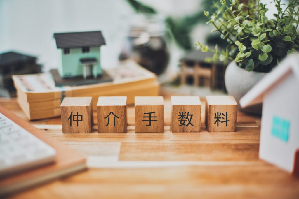Row of wooden blocks displaying the Japanese term 仲介手数料 (brokerage fee) on a desk with office props in the background.