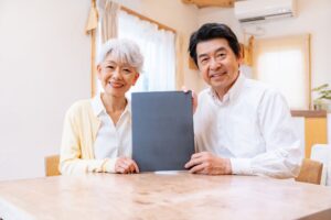 Smiling elderly couple seated at a table, presenting a dark folder toward the camera in a bright home setting at a kitchen/dining area.