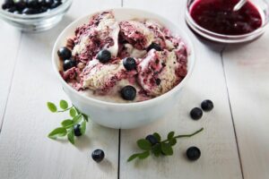 Bowl of blueberry-vanilla ice cream swirls with fresh blueberries on a white wooden table.