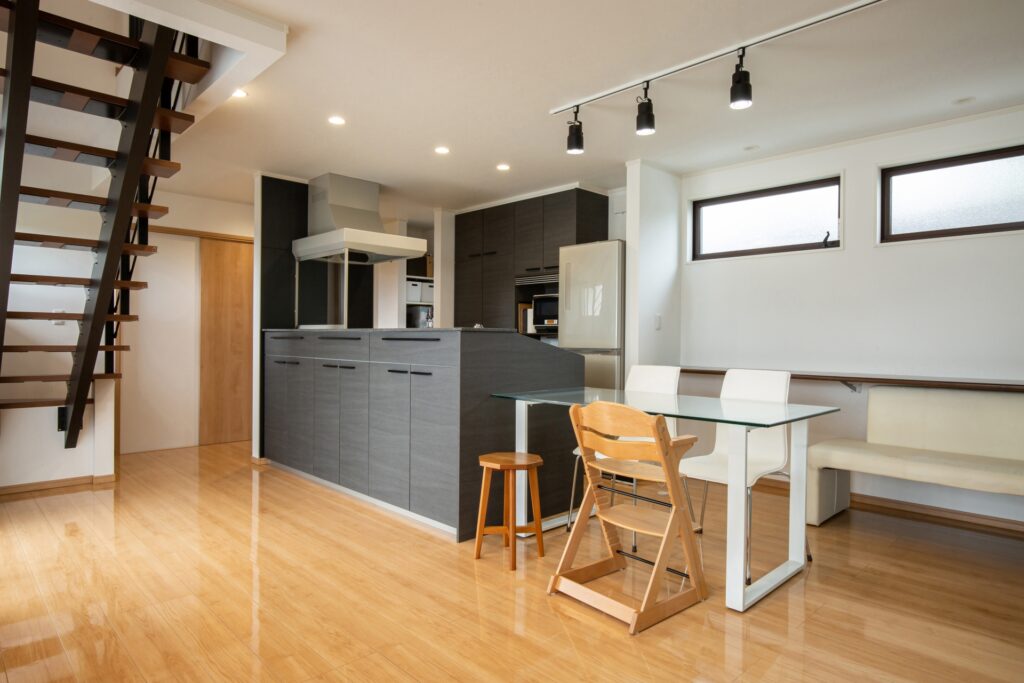 Open-plan kitchen with dark gray cabinets, island, stainless range hood, glass dining table, wooden chairs, and a wooden staircase on the left.