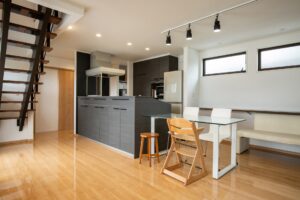Open-plan kitchen with dark gray cabinets, island, stainless range hood, glass dining table, wooden chairs, and a wooden staircase on the left.