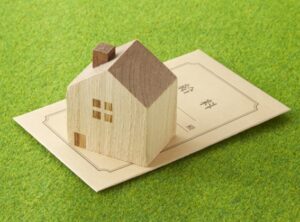 Wooden house model resting on a formal document, symbolizing real estate or home ownership on green grass.