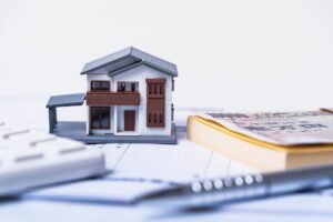 Model house placed on financial documents and money, illustrating real estate financing. (Toy house with papers and currency on a desk)