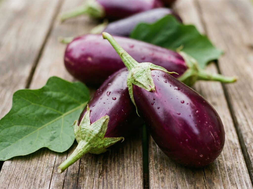 Purple eggplants with green stems and leaves resting on a weathered wooden table. Water droplets cling to the skins.]"