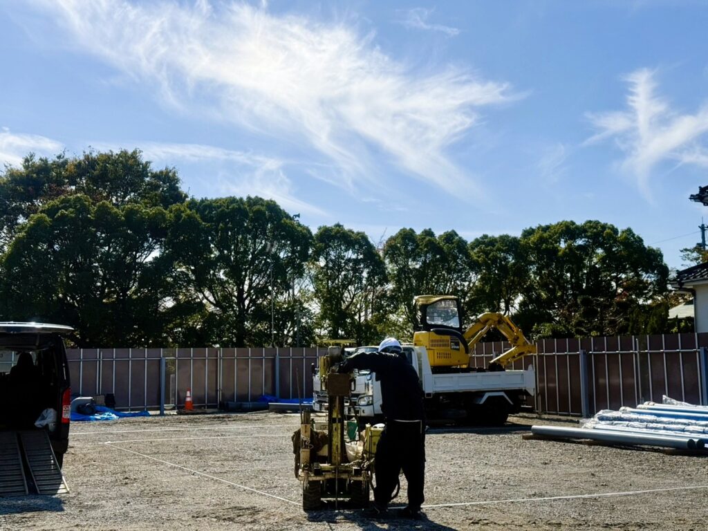 Construction site with a worker wearing a helmet operating a cart, a yellow mini excavator on a truck, and a chain-link fence under a blue sky.