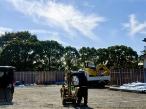 Construction site with a worker wearing a helmet operating a cart, a yellow mini excavator on a truck, and a chain-link fence under a blue sky.